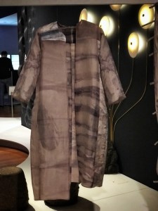 Photo from the exhibit of an open-front silk coat dyed using Chieza's process. It is a brown grey with both horizontal and vertical seepage shaped streaks of a dull purple. The coat has no buttons, collar or cuffs. 