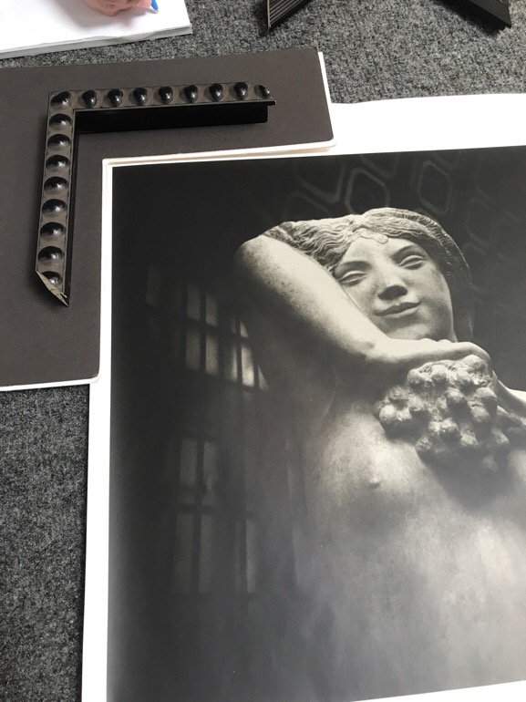 A black and white photograph of a stone sculpture of a nude woman holding grapes with a frame sample that has raised grape shapes on the border