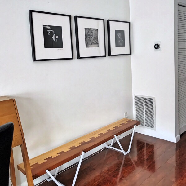 Black framed black and white photos on a white wall above a modern bench with a dovetail plank seat and white metal legs