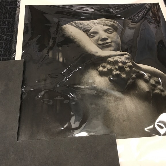 Black and white photograph of a stone sculture of a nude woman holding grapes framed by a sample of black matte board