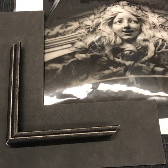 Black and white photograph of a stone sculpture of a young woman's head framed by a sample of matte board and a sample of a silvery black frame.
