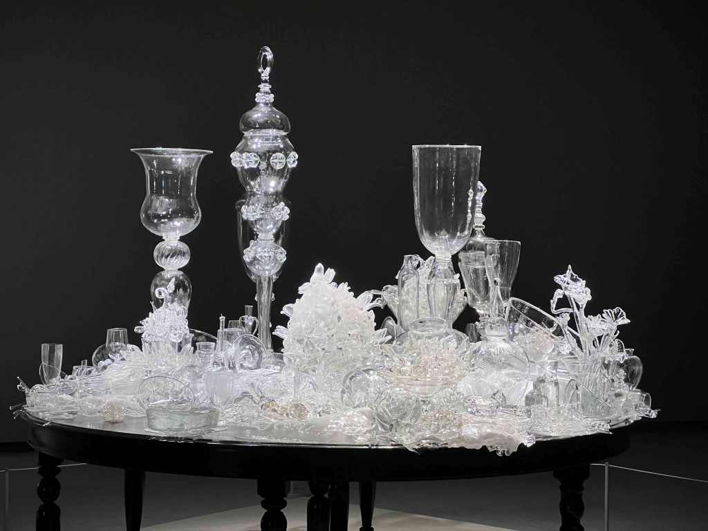 ornate clear blass pitchers, vases, glasses, flowers sit on tip of a round black table