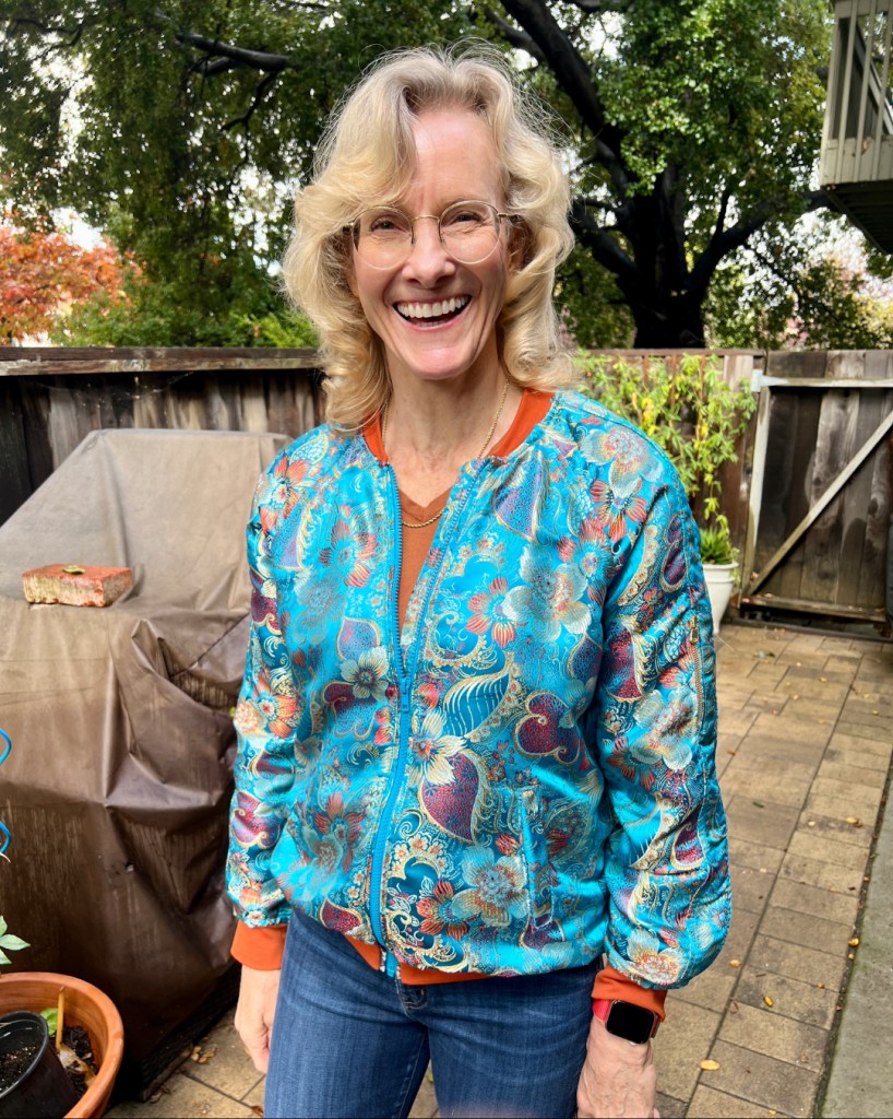 A smiling person wearing a colorful turquoise brocade bomber jacket with ribbed orange accents, standing outdoors in a relaxed setting.