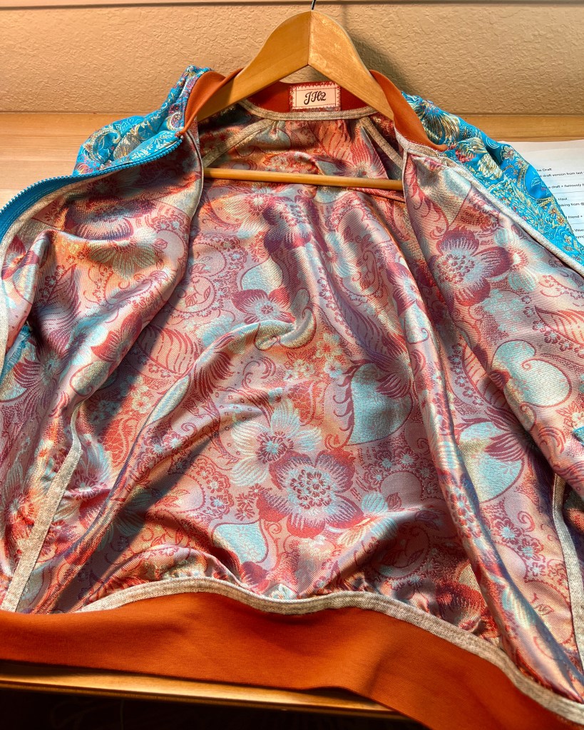 The interior view of a bomber jacket featuring a vibrant brocade lining with floral patterns in shades of orange/red, complemented by a cinnamon ribbing at the waist.